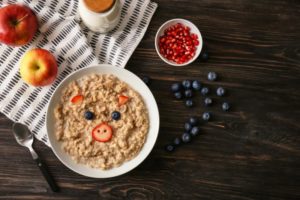 Bowl of oatmeal with a face made of berries. The bowl is surrounded by apples, blueberries, and diced strawberries.