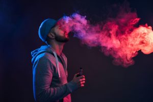 Man wearing a hoodie and baseball cap smoking a vape in red and purple lighting