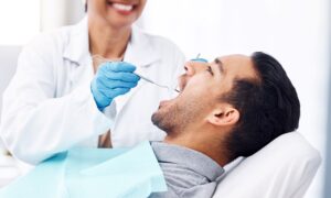 Man with dark hair and grey shirt having oral examination done by dentist with blue gloves