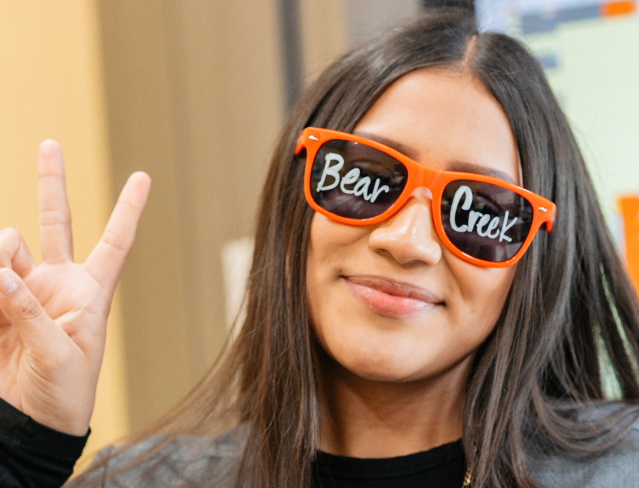 Woman with Bear Creek written on her orange sunglasses smiling and doing peace sign